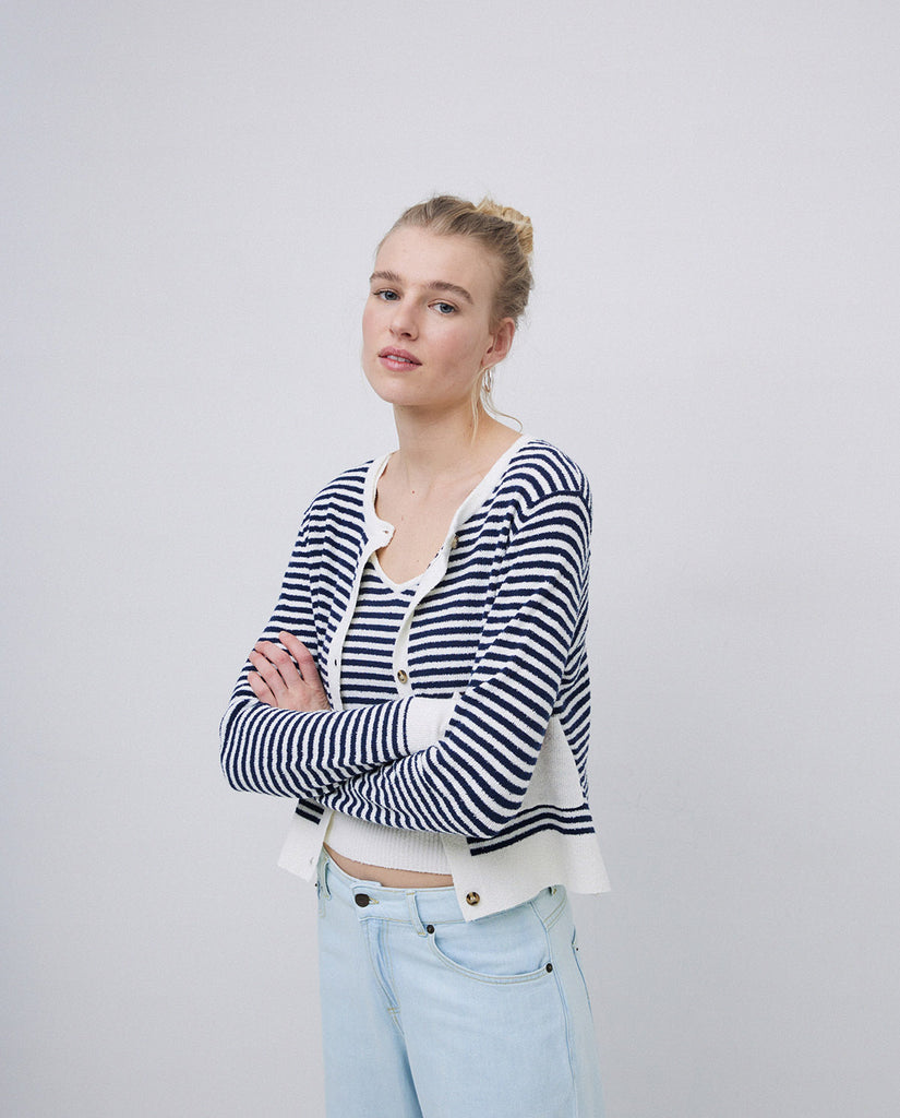 A woman with her arms folded  wearing a navy and white stripped cardigan white white edges, two front pockets and round neckline. 