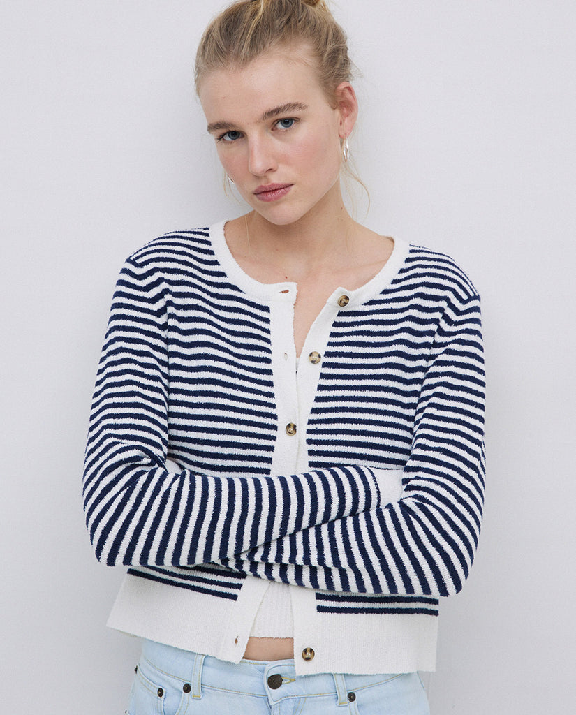 A woman with her arms folded  wearing a navy and white stripped cardigan white white edges, two front pockets and round neckline. 