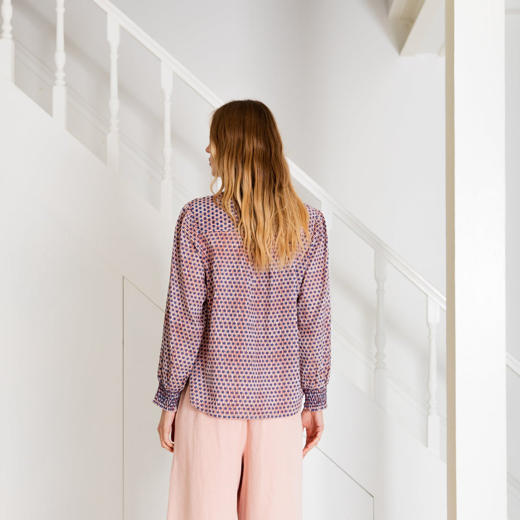 A woman stands in a Bonte top featuring a purple and pink pattern, paired with light pink trousers.