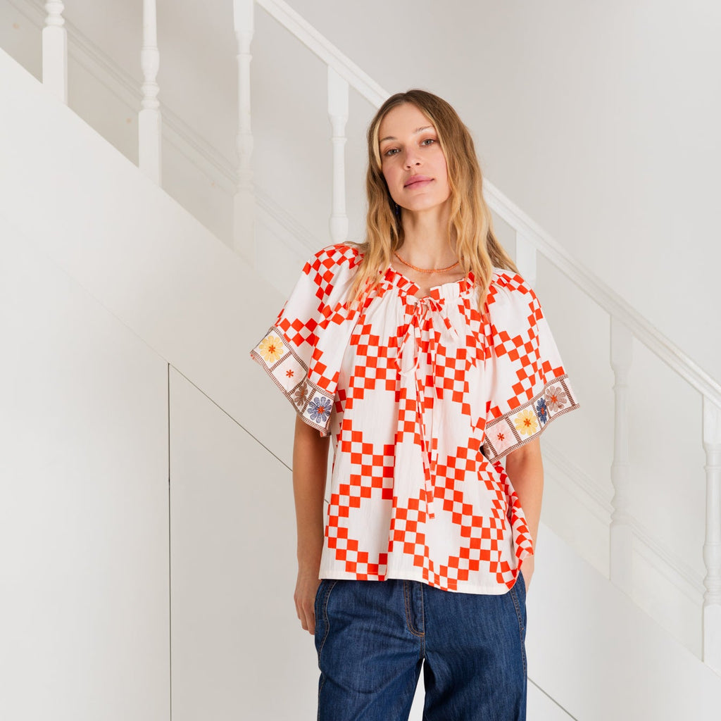 A woman stands in a staircase wearing a Bonte top with red and white checkered patterns and blue, yellow, and orange details.