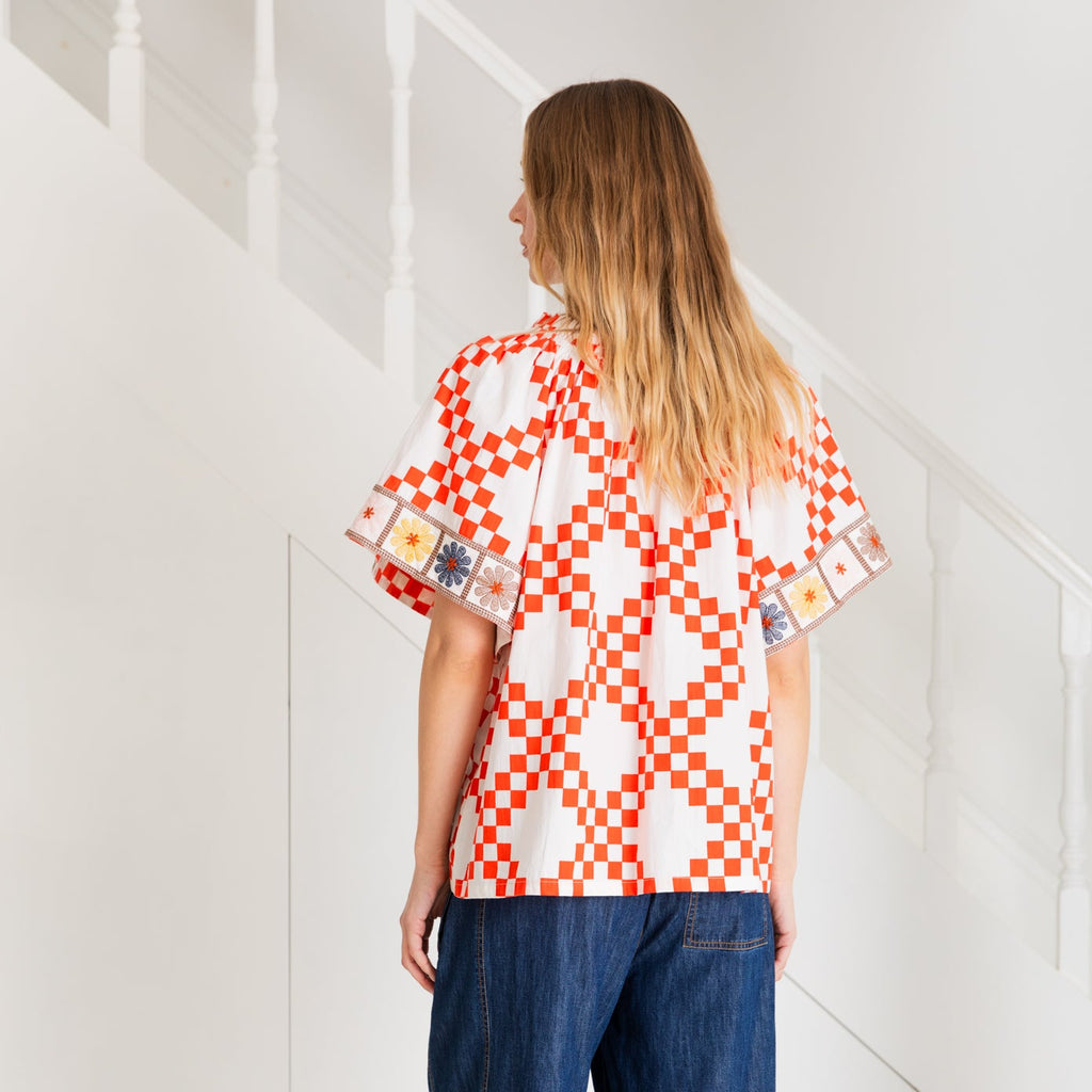 Woman wearing a Bonte top with red and white checkered pattern and colorful embroidered sleeves, standing by stairs.