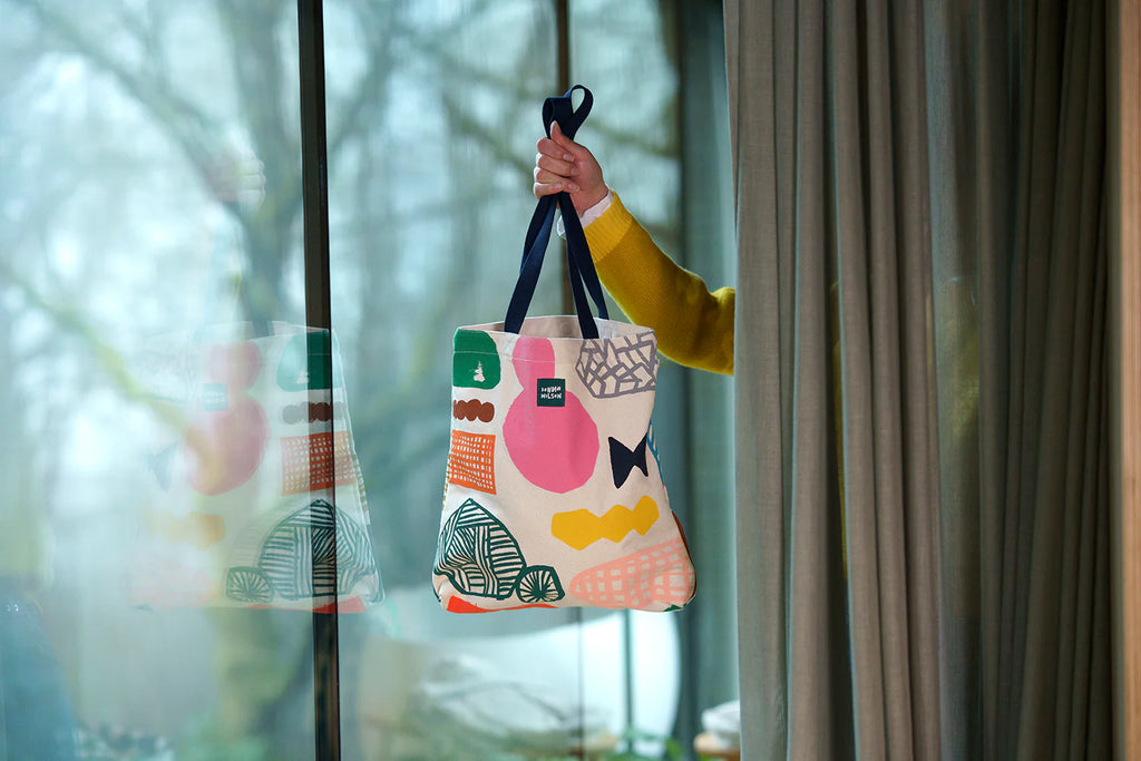 A hand holds a colorful Donna Wilson tote bag with green, pink, orange, and black patterns, against a curtain backdrop.