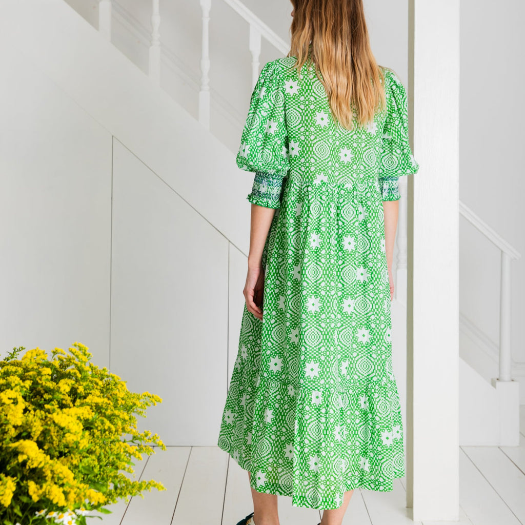 A woman in a green floral dress by Bonte stands against white stairs, with a bouquet of yellow flowers nearby.