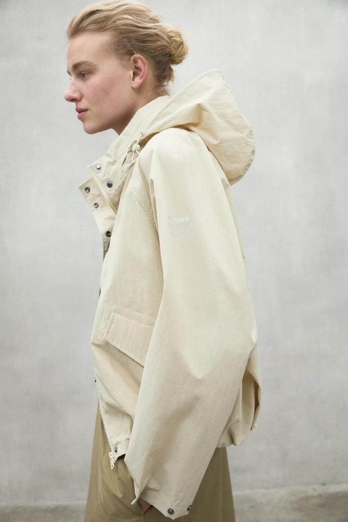 A woman in a beige Ecoalf jacket poses against a gray wall, her hair styled in a bun.