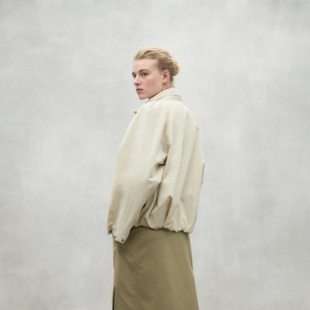 A model in a beige Ecoalf jacket and a khaki skirt poses against a gray background, showcasing a stylish look.