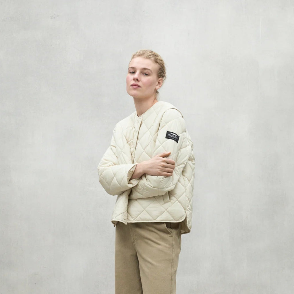 A woman poses in a cream Ecoalf quilted jacket and beige pants against a gray background.