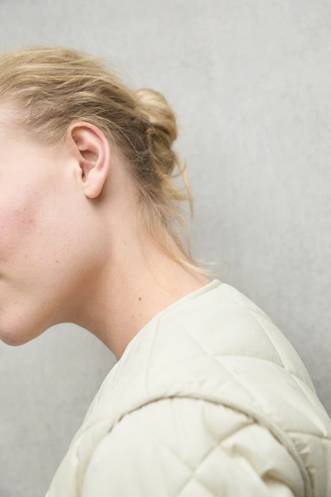 A close-up of a woman wearing a light cream Ecoalf quilted jacket, with her hair tied back.