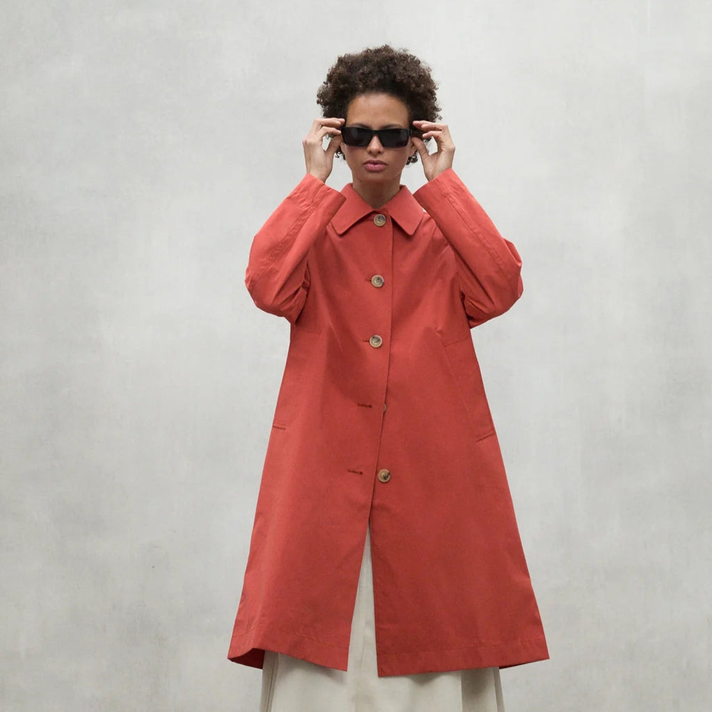 A model in an Ecoalf coral coat with wooden buttons, wearing sunglasses, posed against a neutral background.