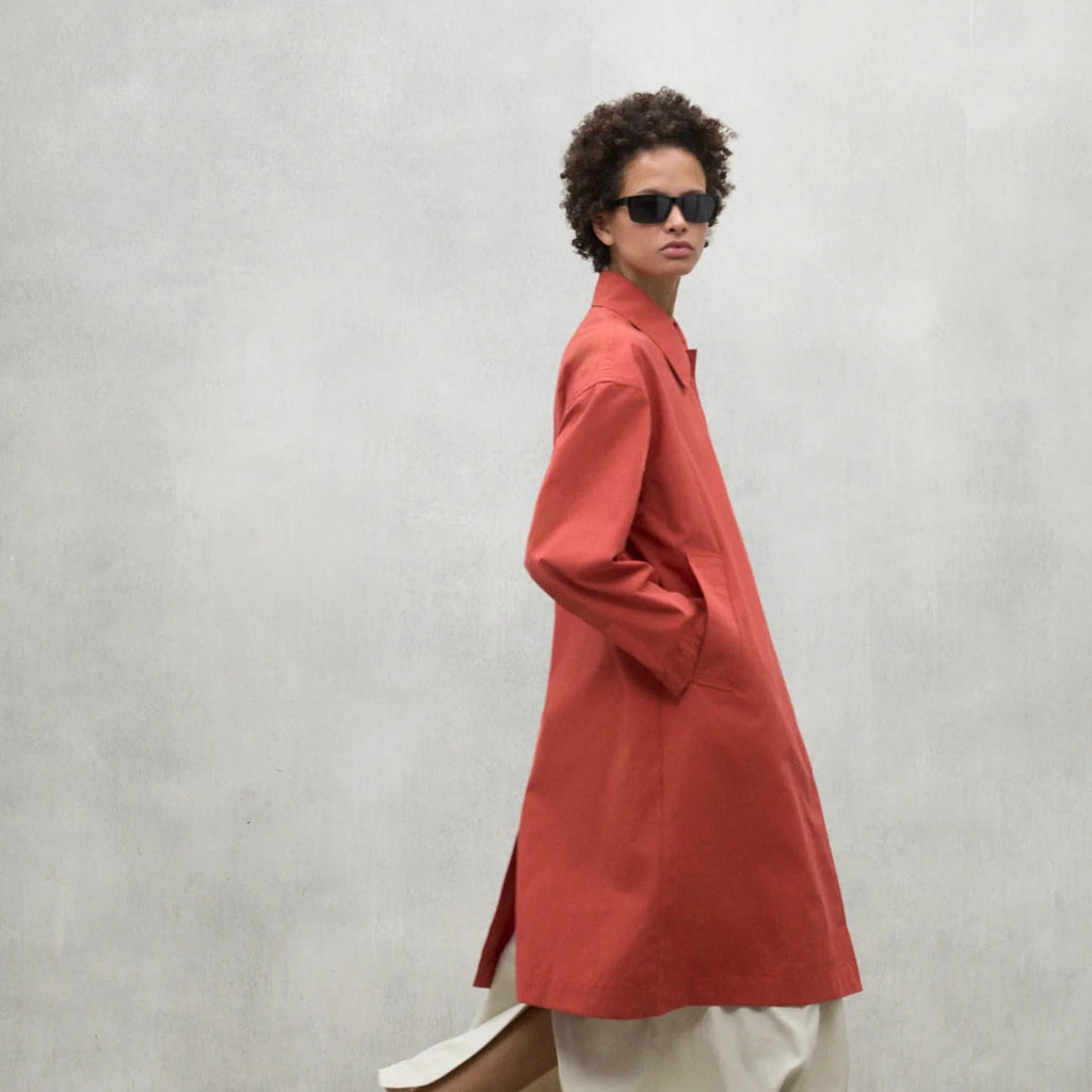 A model wears a coral Ecoalf coat with pockets and black sunglasses, paired with a light cream skirt against a grey backdrop.