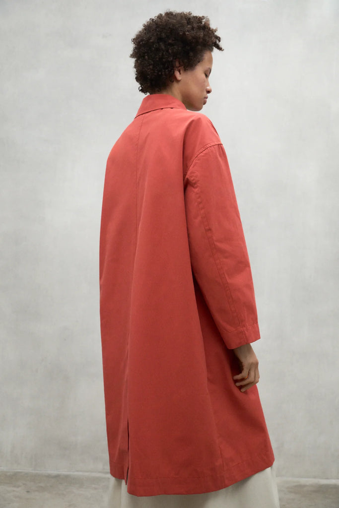 A person wearing a coral Ecoalf coat stands against a gray wall, showcasing a minimalist style.