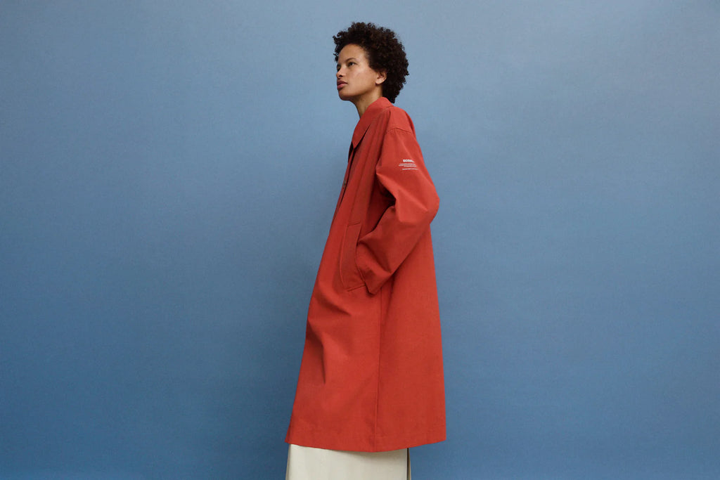 Model in a red Ecoalf coat against a blue background, with a white skirt peeking out below.