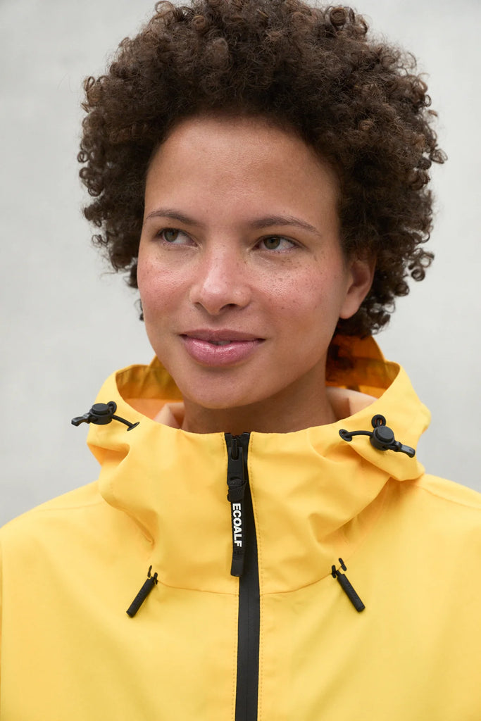 A close-up of a person wearing a bright yellow Ecoalf jacket with a black zipper and details, smiling softly.