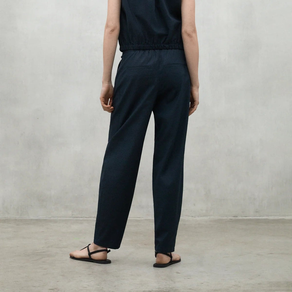 A person stands with their back to the camera, wearing a navy Ecoalf jumpsuit and black sandals on a concrete floor.