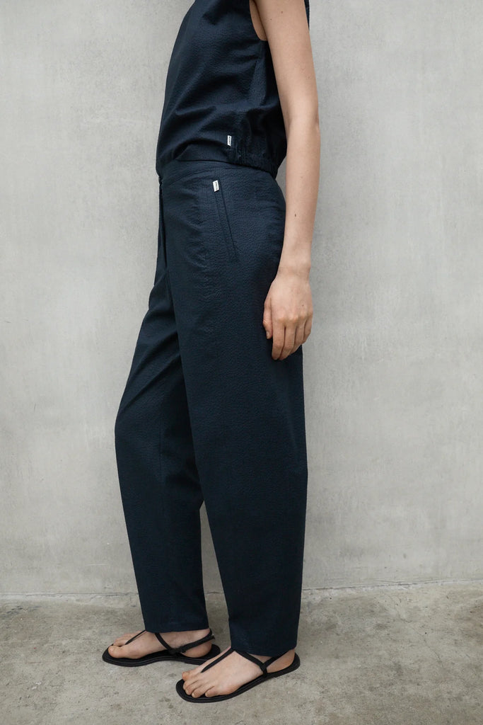 Model in Ecoalf dark navy outfit stands against a gray wall, wearing black sandals.