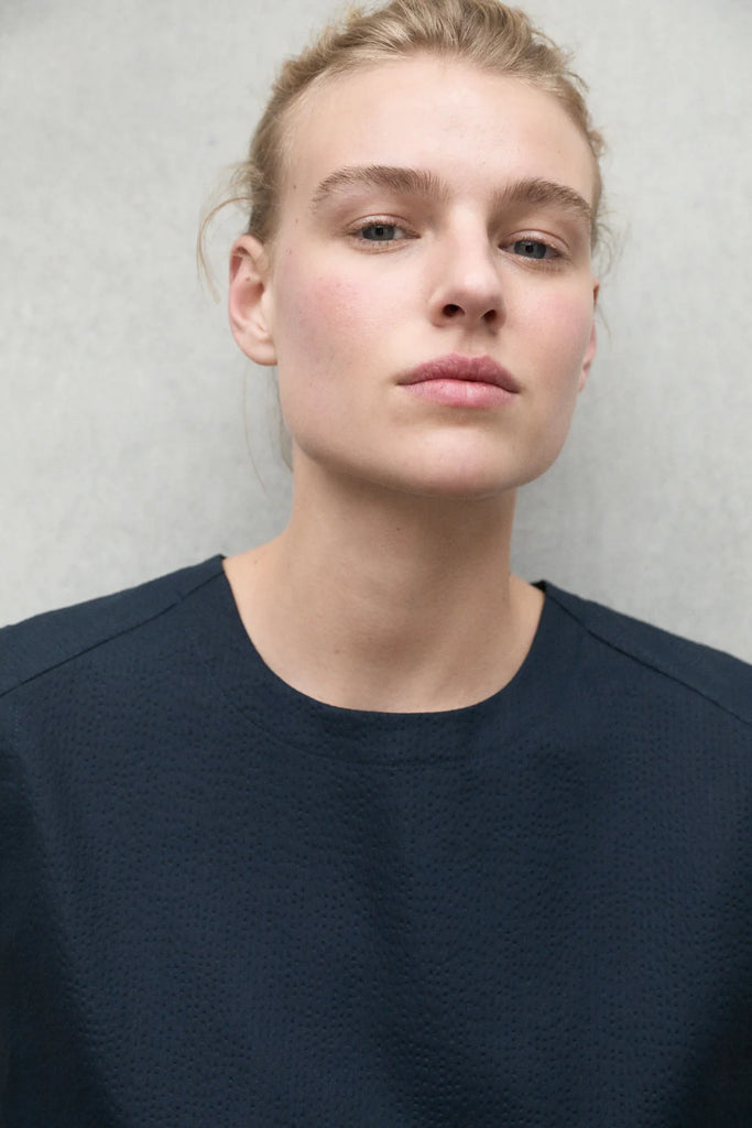 Close-up of a model wearing a dark navy Ecoalf top, with a neutral background and natural makeup.