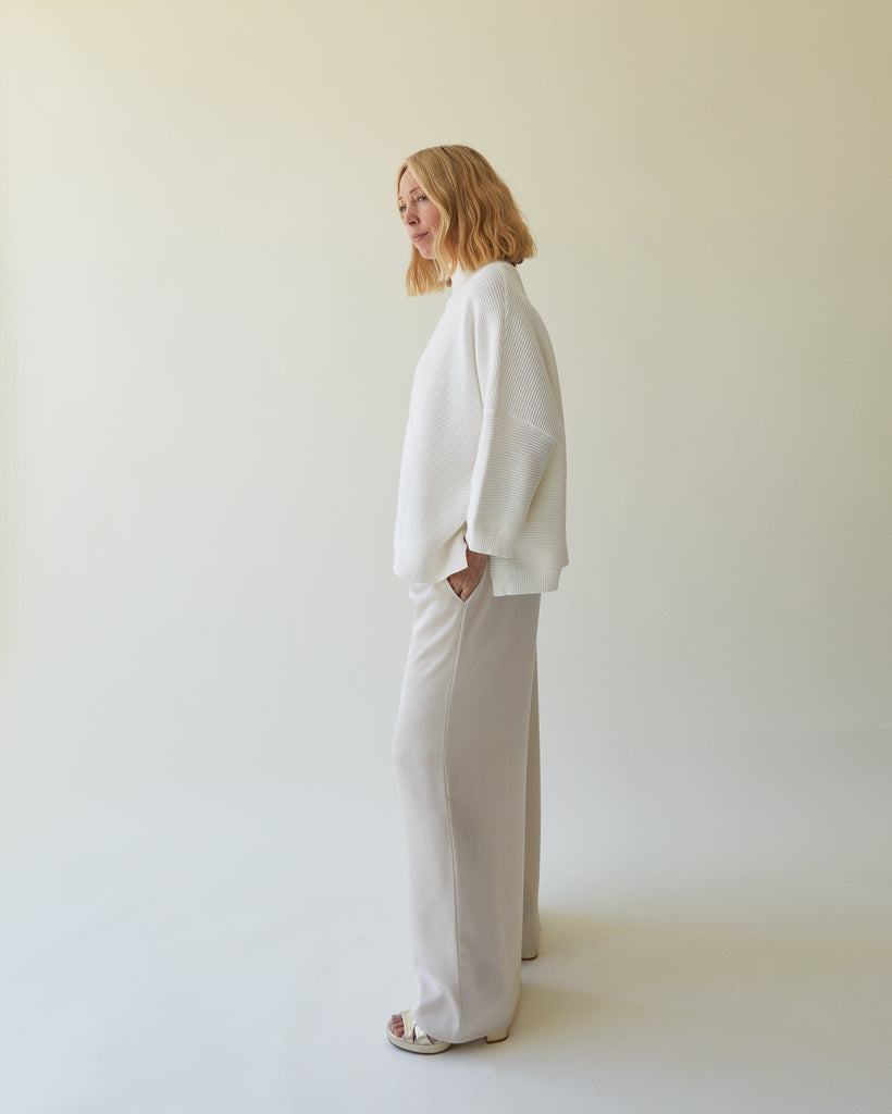 A model in Chalk UK clothing, wearing a white sweater and light beige pants, stands against a neutral background.