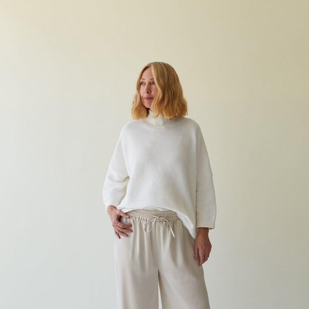 A woman wearing a white top and beige pants from Chalk UK, standing in front of a light background.